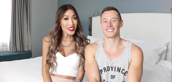 Two friends - a gay youtuber in a tank top and a trans woman in a white vest top - sit together on a bed.