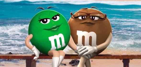 The green M&M (left) sitting on a bench on the beach with her lesbian partner, the brown M&M