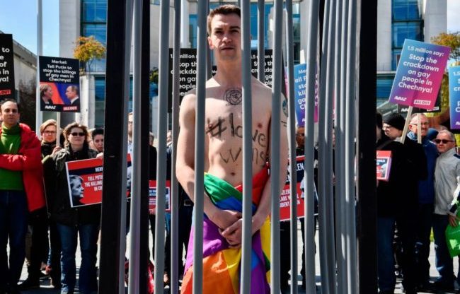 An activist stands naked, wrapped in a rainbow flag, in a mock cage