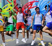 A group of men in white vests and shorts. Each holds balloons in a different colour of the rainbow.