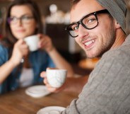 A man smiles at the camera while his girlfriend drinks coffee