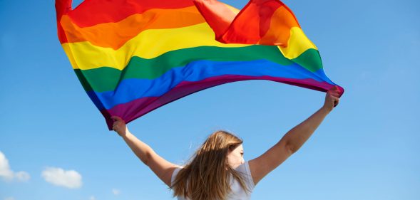 Woman waving an LGBT+ Pride flag against a blue sky