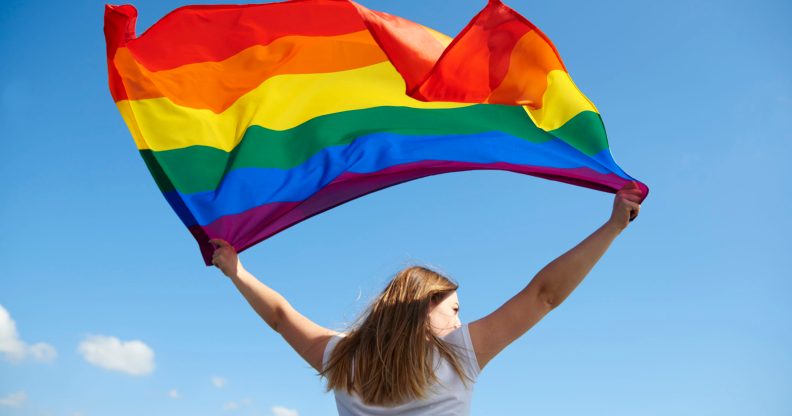 Woman waving an LGBT+ Pride flag against a blue sky