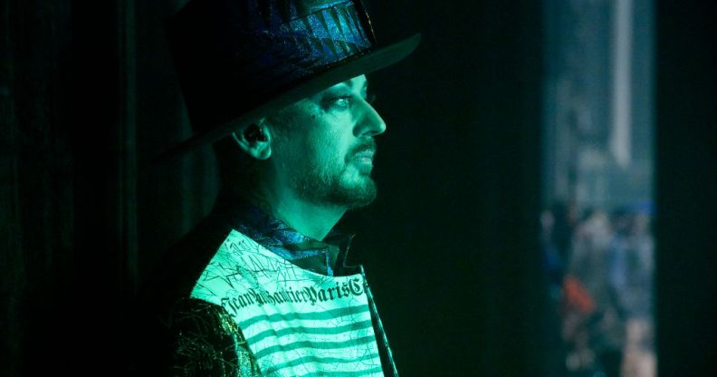 The singer Boy George stands side-on wearing a hat with a green light shining on him