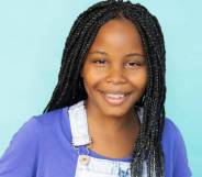 AJ Nesbitt smiling, wearing denim dungarees and a purple t-shirt, her hair in long braids