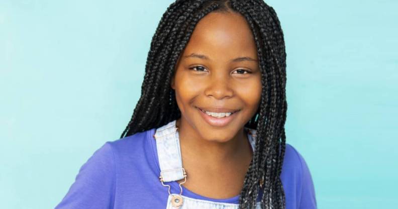 AJ Nesbitt smiling, wearing denim dungarees and a purple t-shirt, her hair in long braids