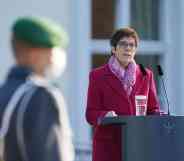 Annegret Kramp-Karrenbauer stands at a podium in a coat. A blurred serviceman is seen in the foreground.