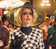 Anne Hathaway in The Witches, wearing a big black hat, blonde hair, and carrying a black cat in a grand ballroom