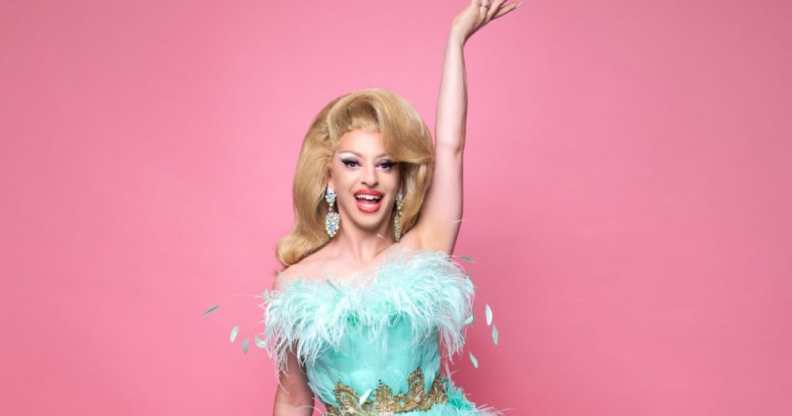 Miz Cracker in a fluffy blue gown against a pink background, with one hand in the air, smiling