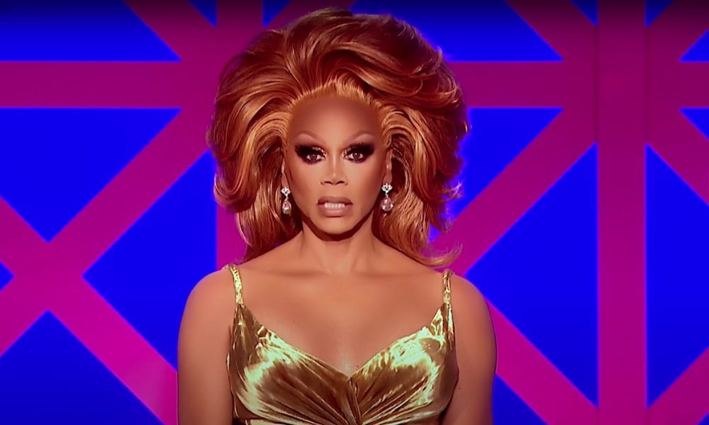 RuPaul in a gold dress with copper hair sitting in front of the Drag Race UK judges' panel backdrop