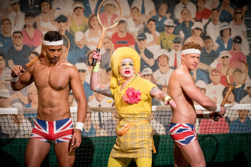 Ginny Lemon in a yellow suit holding a tennis racket, flanked by two Brit crew wearing just Union Jack boxers