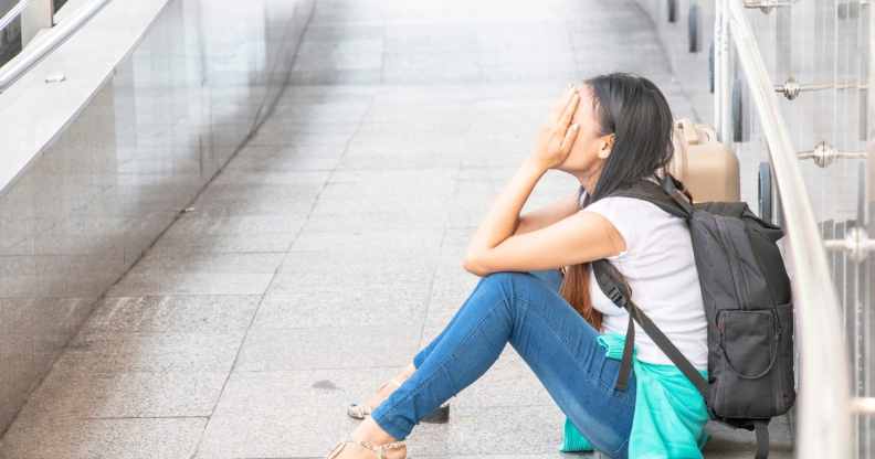 A woman sits on the floor with her hands on her face, crying, while wearing a backpack