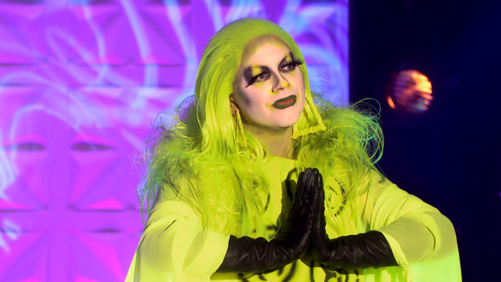 Ginny with her hands together in a prayer motion, wearing a highlighter yellow dress and matching hair