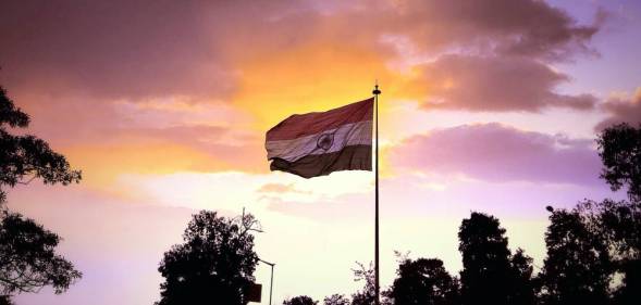 A photo of the national flag of India showing trees in the background and a pink/orange sunset sky