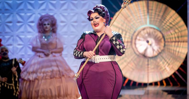 Lawrence Chaney, a drag queen in a long purpler dress with racing flag tripe down the sides, holding a sceptre