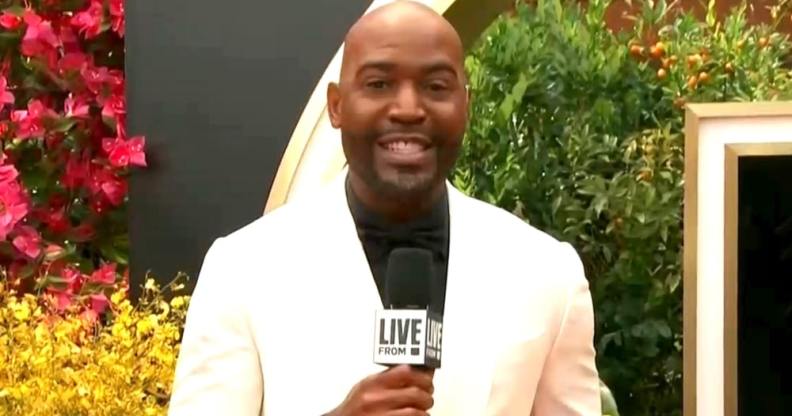 Karamo Brown holds a microphone in a white blazer and black shirt on the Oscars red carpet