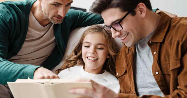 Happy gay parents reading book together with their little daughter at home