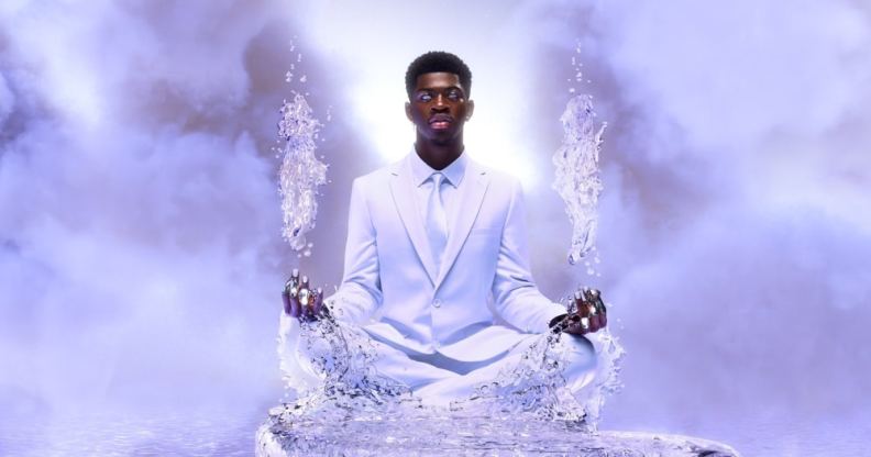 Lil Nas X sits in a white suit on a pathway made of ice floating in the skies