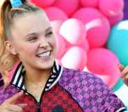 JoJo Siwa stands in front of colourful balloons while wearing red and purple jacket