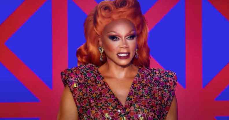 RuPaul appears in a red wig and red and blue outfit in front of a geometric patterned background