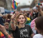 Young person holds up an LGBT+ pride flag while wearing a shirt that says 'love is love'