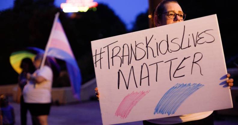 An image shows a trans rights advocate holding a sign that reads 'trans kids lives matter'