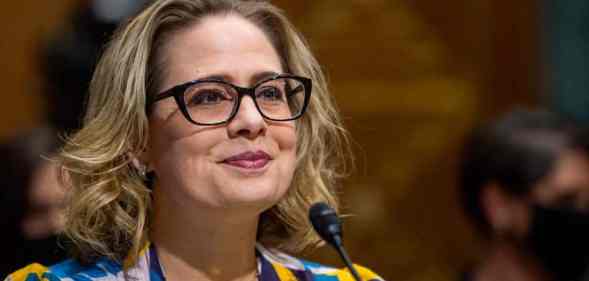 Kyrsten Sinema wears a black, blue, yellow and white patterned top while speaking into a microphone at a US Senate Committee on Finance hearing