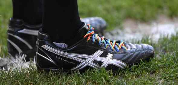 A person wearing black trainers stands on a football pitch. The trainers have rainbow laces to show support for the LGBT+ community