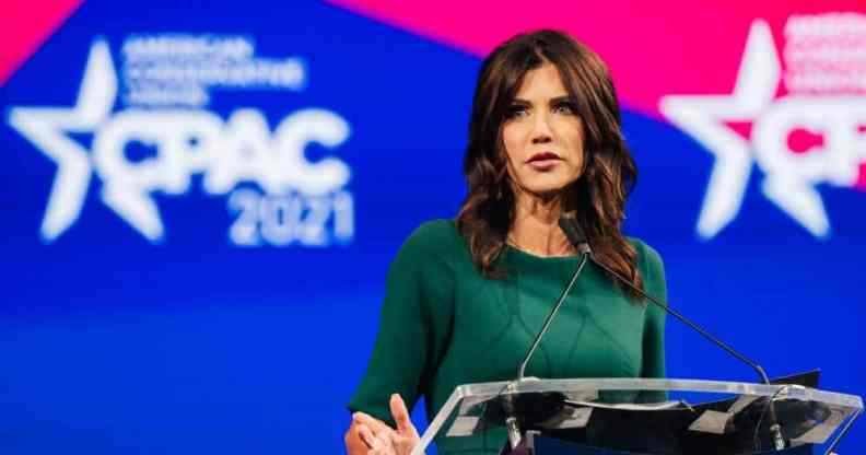 Kristi Noem wears a green outfit and stands at a podium in front of a red and blue background