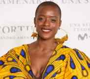 T'Nia Miller smiles at the camera while wearing a beautiful yellow, brown and blue dress and gold hoop earrings