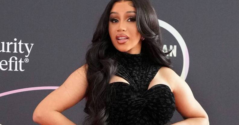 Cardi B wears a black dress with a neck piece amid a dark background