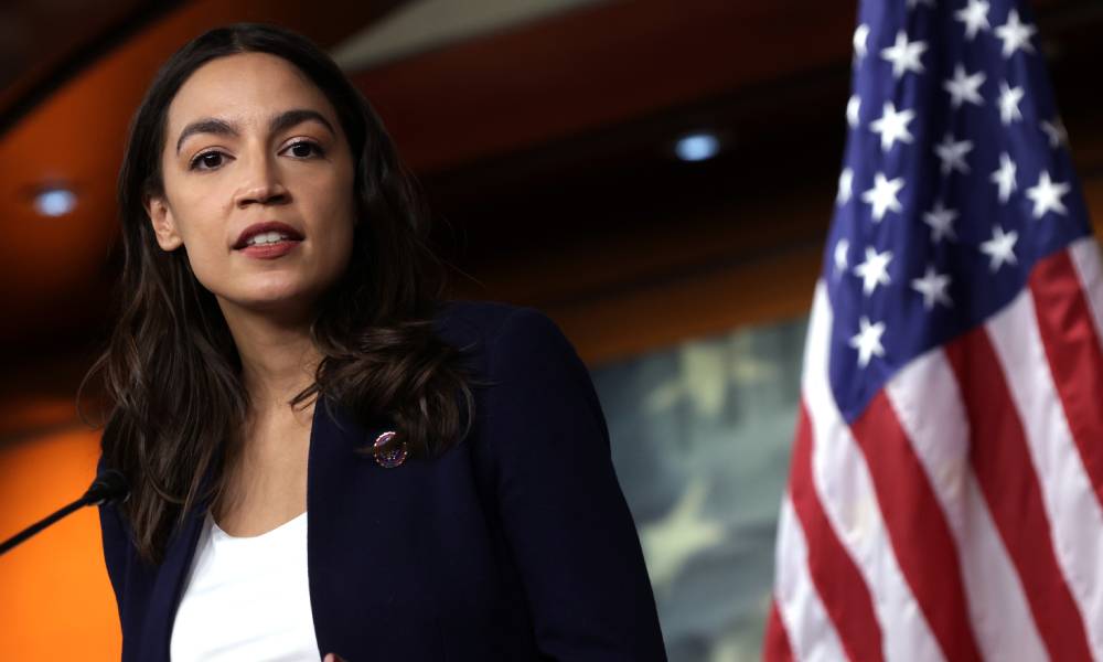 Alexandria Ocasio-Cortez looks on at the photographer while wearing a black jacket and white top. She is speaking at a podium with a US flag in the background