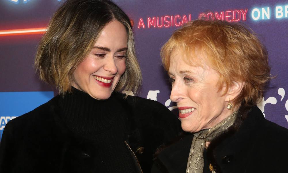 Sarah Paulson and Holland Taylor smile at each other. Both women are dressed in black, and Taylor has a lighter grey scarf around her neck