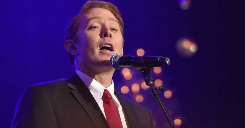 Clay Aiken sings into a microphone as he wears a white shirt, black jacket and red tie