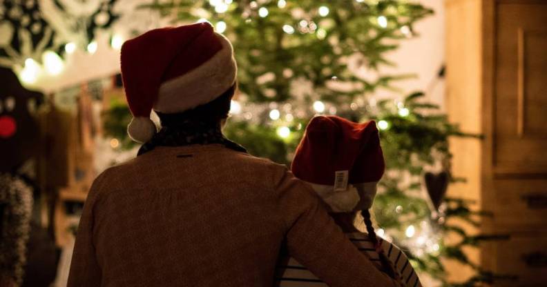 an older person wraps their arm around a younger person. Both are wearing Christmas hats as they look on at a Christmas tree