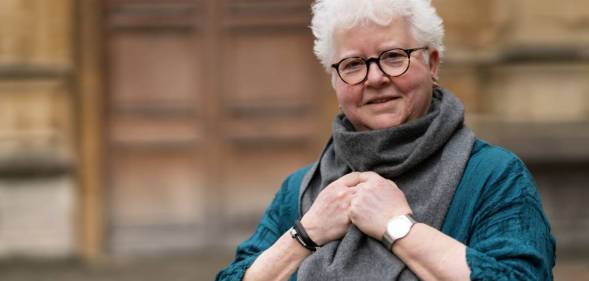 Val McDermid, a white woman, wears a blue shirt and a grey scarf that her hands are clutching onto