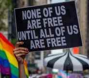 A participant holds a sign reading "None of us are free until all of us are free" and an LGBT+ Pride flag can be seen behind during the Reclaim Pride Coalition's third annual Queer Liberation March