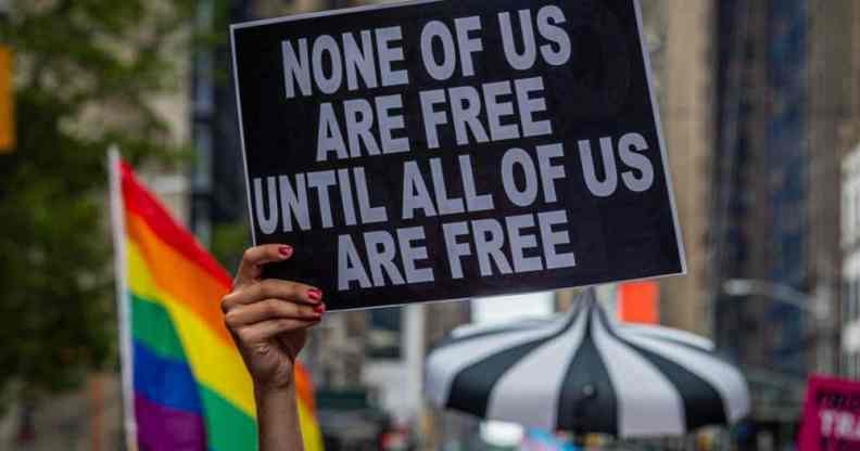 A participant holds a sign reading "None of us are free until all of us are free" and an LGBT+ Pride flag can be seen behind during the Reclaim Pride Coalition's third annual Queer Liberation March