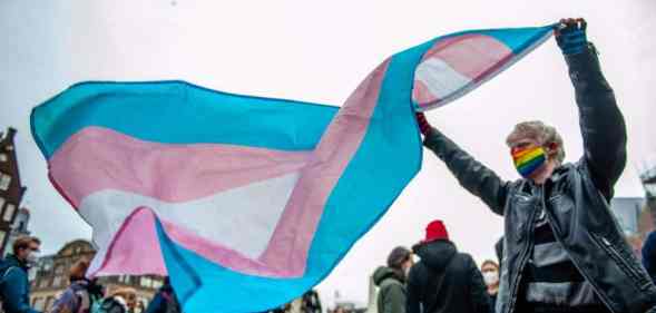 A person wearing a rainbow coloured face mask holds up a transgender Pride flag above their head