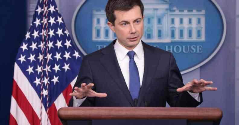 Pete Buttigieg stands at a podium dressed in a suit and tie with his hands raised