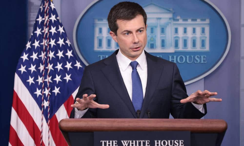 Pete Buttigieg stands at a podium dressed in a suit and tie with his hands raised