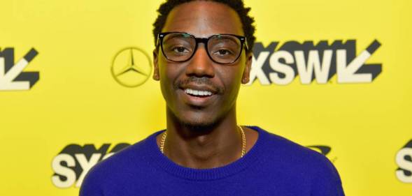 Jerrod Carmichael smiles at the camera while wearing a blue-purple top in front of a bright yellow background