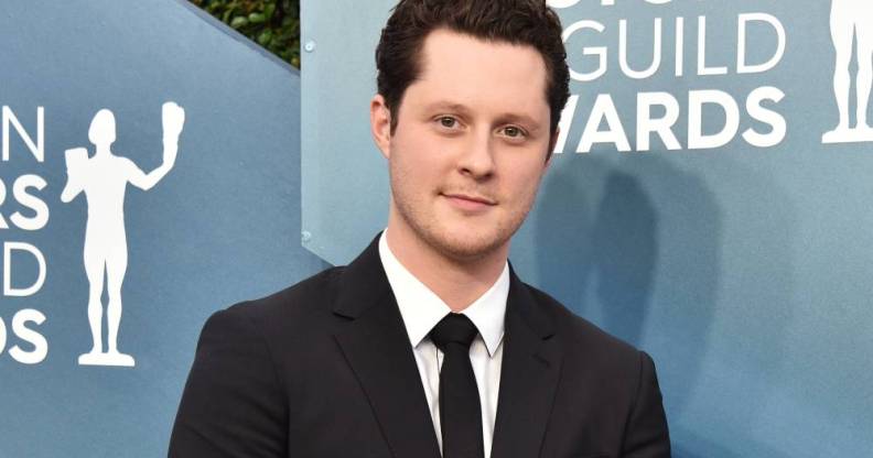 Schitt's Creek star Noah Reid smiles for the camera while wearing a suit and standing in front of a blue background