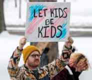 A person holds up a sign reading "let kids be kids" in front of the colours of the transgender pride flag