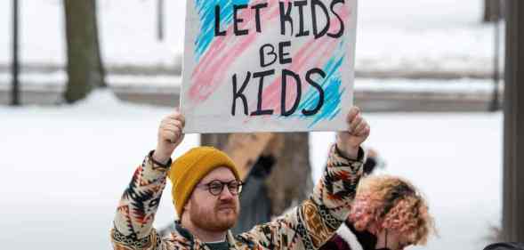 A person holds up a sign reading "let kids be kids" in front of the colours of the transgender pride flag