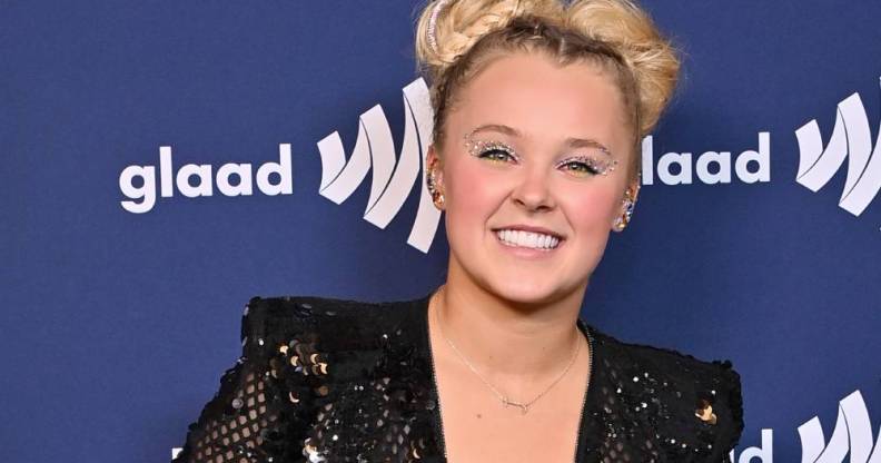 JoJo Siwa smiles at the camera as she wears a black sequinned outfit and stands in front of a dark blue background with the white GLAAD logo on it