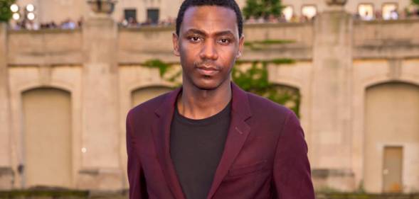 Jerrod Carmichael stares at the camera while wearing a black shirt and a dark red jacket