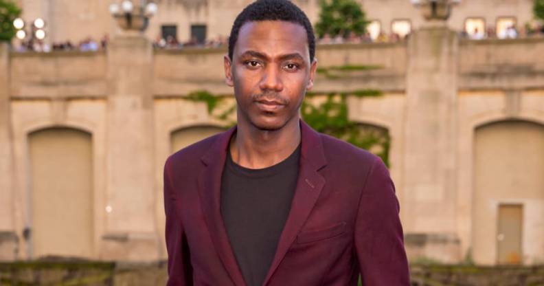 Jerrod Carmichael stares at the camera while wearing a black shirt and a dark red jacket