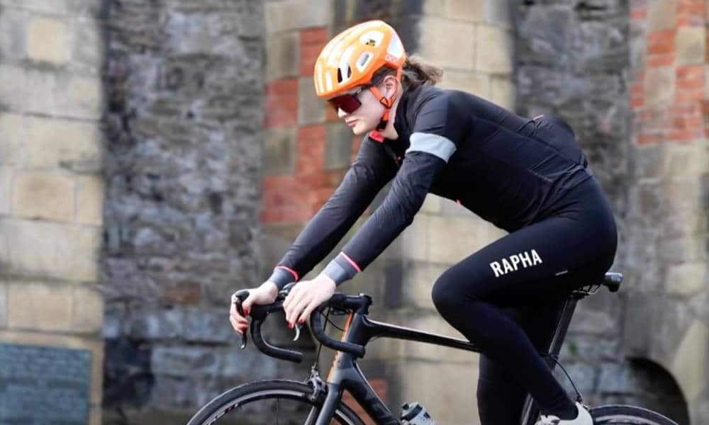 Trans cyclist Emily Bridges rides a black bicycle in front of a brick building