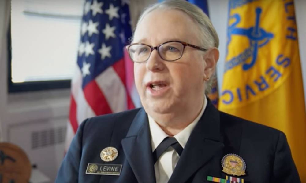 Dr Rachel Levine speaks to a person off camera while wearing a blue uniform with an American flag in the background